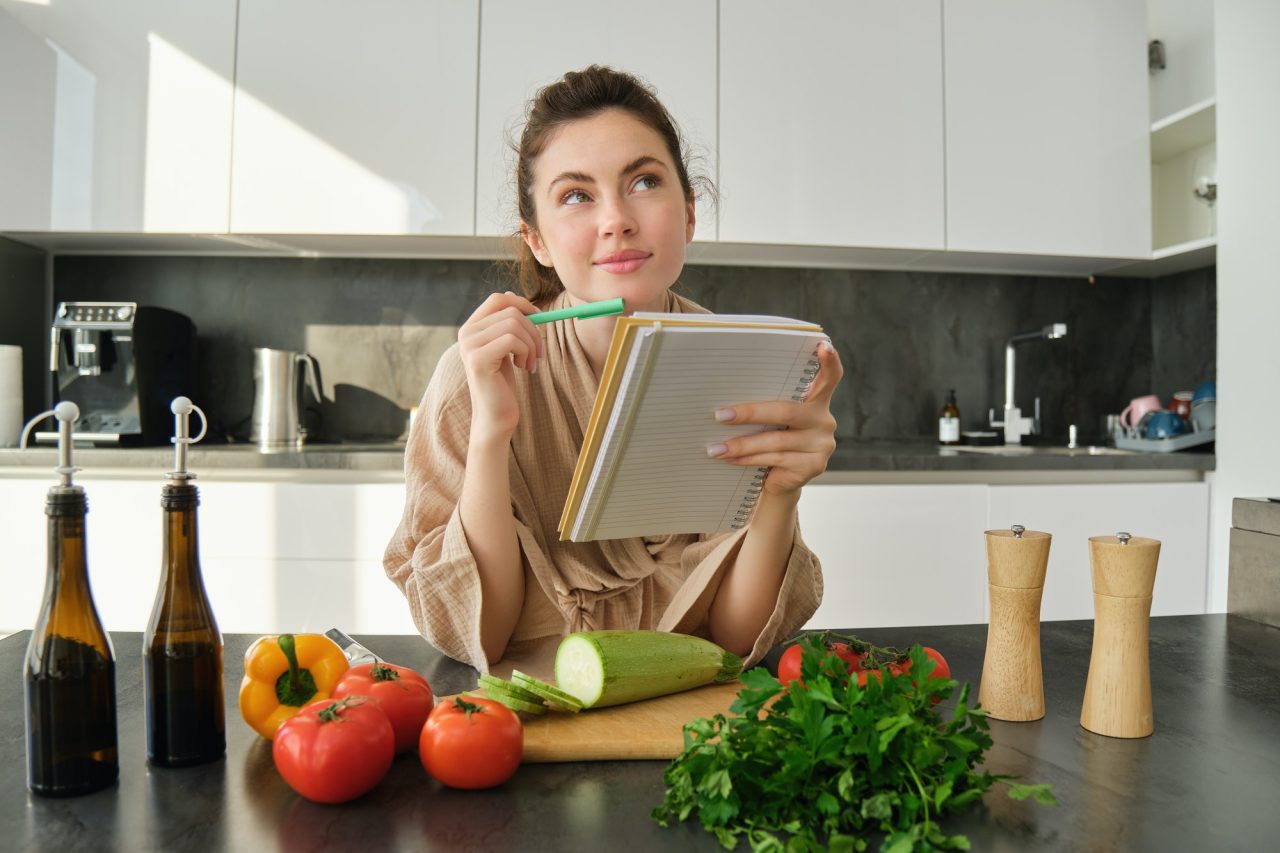 portrait of woman checking recipe notes in notebook standing in kitchen with vegetables cooking.jpg